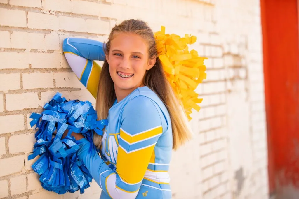 Cheerleader with braces smiling and holding blue and yellow pom-poms against a textured white wall, representing youth sports and school spirit.