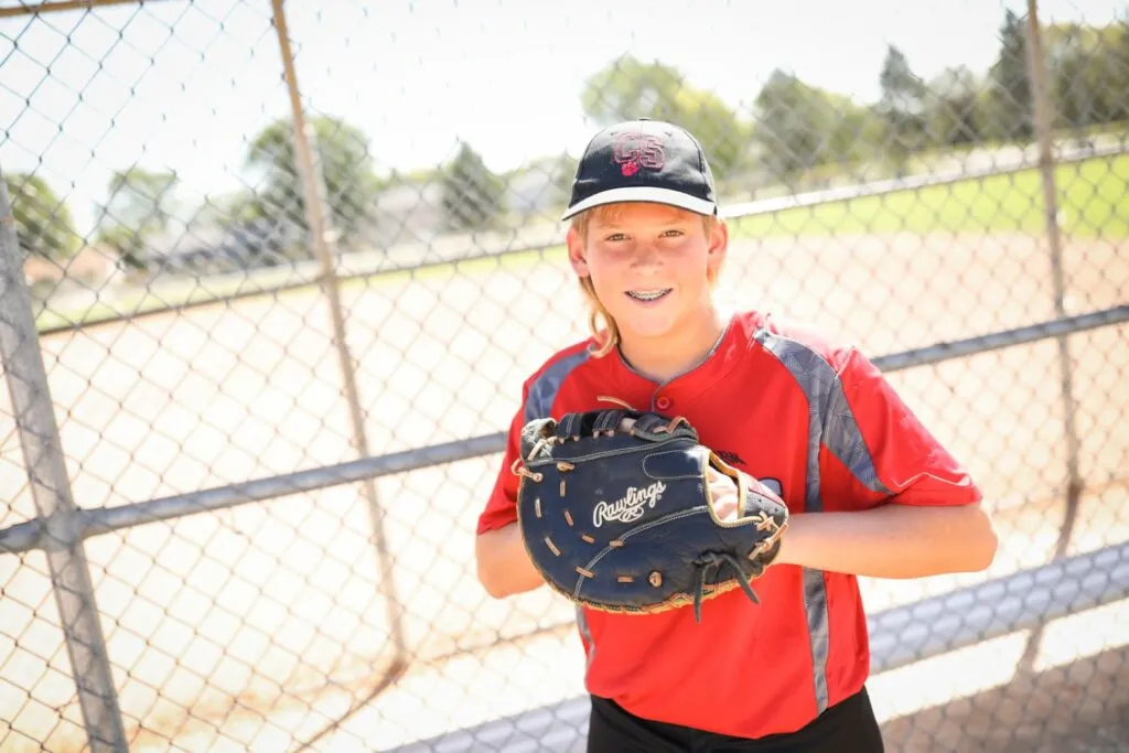 Young boy in red baseball uniform smiling and holding a baseball glove, with a baseball field and fence in the background, representing active lifestyle and youth sports.