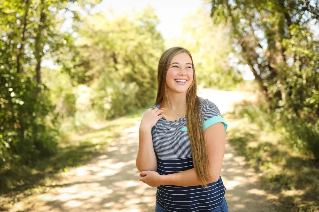 Smiling young woman in casual striped shirt standing on a scenic path surrounded by greenery, reflecting confidence and happiness, relevant to orthodontic care and patient experience at VanLaecken Orthodontics.
