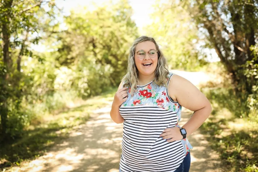 Smiling woman in a floral and striped top standing on a tree-lined path, representing a friendly and approachable atmosphere for orthodontic consultations.