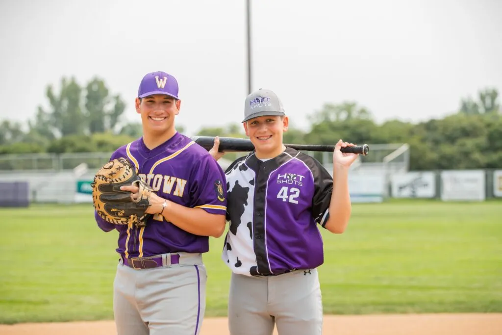 Two young baseball players posing on a field, one wearing a purple jersey with "W" logo and holding a glove, the other in a black and purple jersey holding a bat, both smiling.