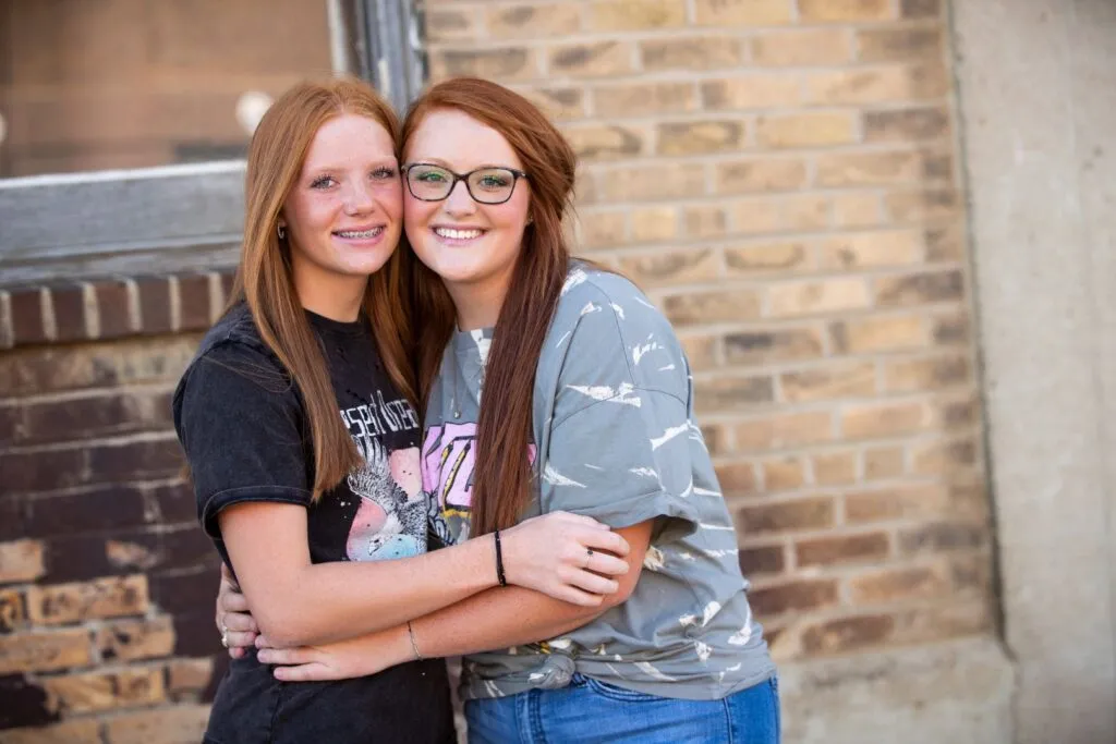 Two smiling teenage girls embracing outdoors, showcasing their braces, against a brick wall, representing the positive experience of orthodontic treatment at VanLaecken Orthodontics.