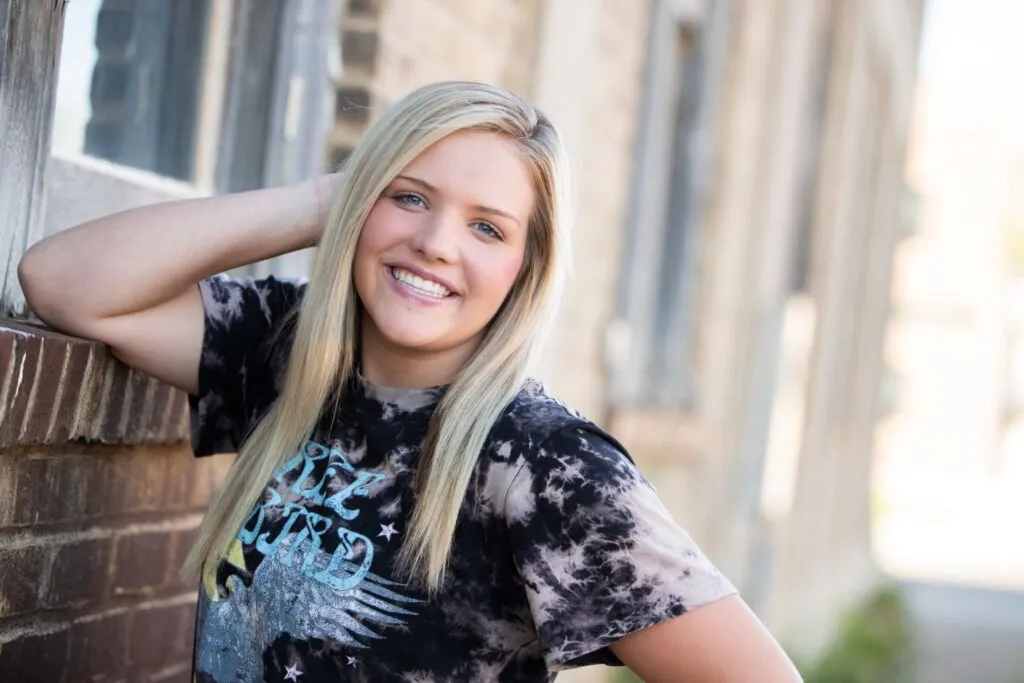 Teen girl smiling confidently against a brick wall, showcasing her straightened teeth, representing the benefits of Invisalign Teen for orthodontic treatment.